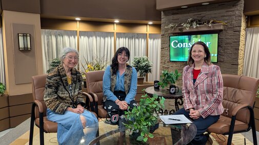 Three people sit in a TV studio with plants, chairs, and a screen displaying "Consider This..."