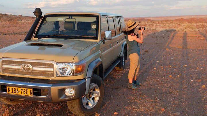 Woman photographing the desert near a parked beige SUV at sunset.