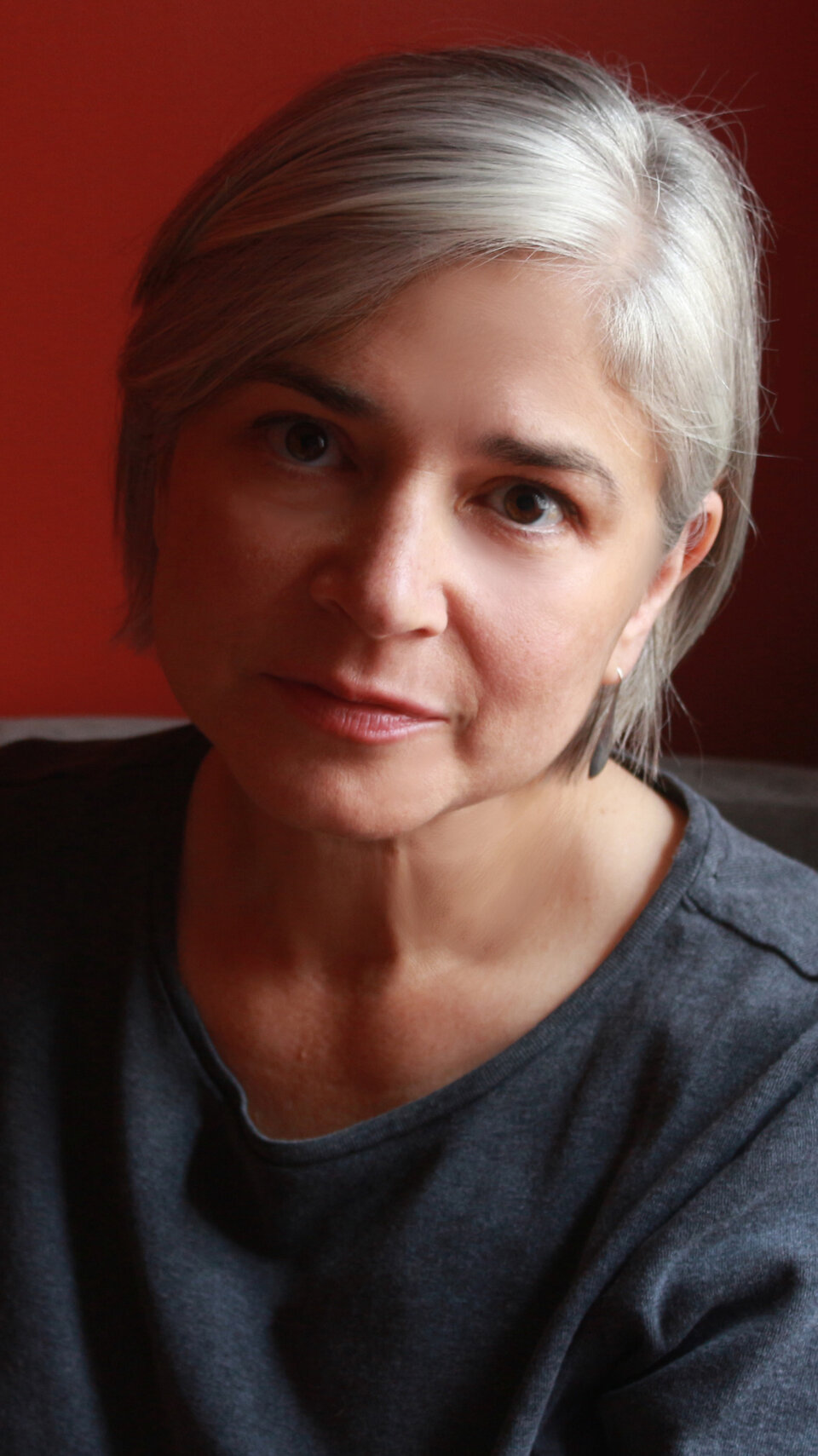 Woman with short gray hair and a neutral expression, wearing a dark top against a red background.